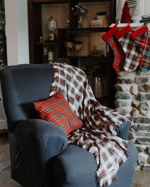 A cozy living room scene featuring a dark gray armchair with a red, white, and black plaid throw blanket draped over it and a matching plaid accent pillow. In the background, a rustic stone fireplace displays red and plaid stockings.