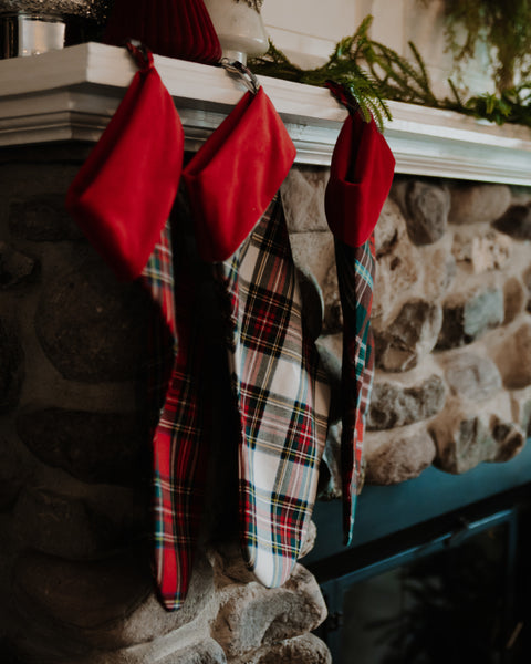 Three Christmas stockings hang from a white fireplace mantel in a rustic stone fireplace. Two stockings are red velvet with red, green, and white plaid bodies, and one is a red and green tartan pattern.