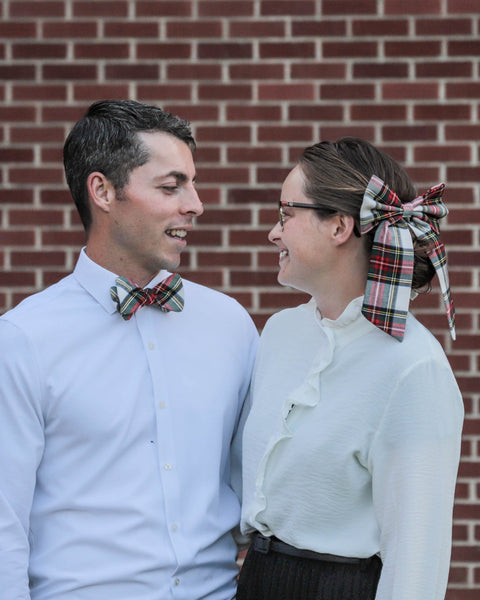 Two people in formal attire, man with plaid bow tie, woman with plaid hair bow with standing in front of a brick wall.