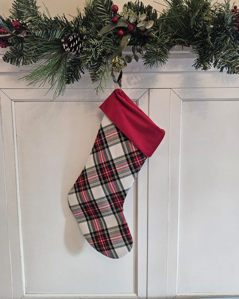 A Christmas stocking with a red and green plaid body and a solid red cuff, hanging on a white paneled wall beneath a green pine garland. Dress Stewart plaid Christmas stocking
