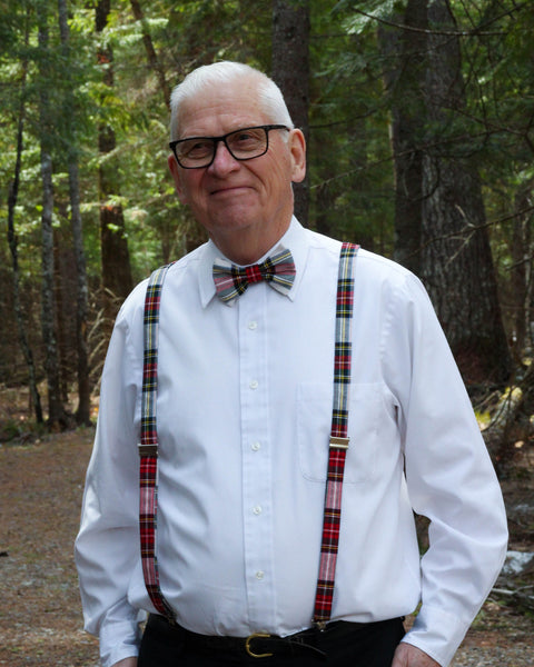 Man wearing a white shirt, white red green and yellow Dress Stewart plaid bow tie, and suspenders standing in a forest.