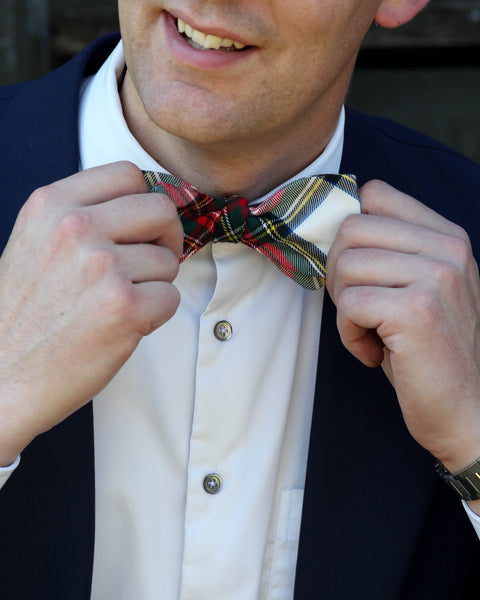 Close up of man adjusting a red and white plaid bow tie with a dark suit and white shirt.