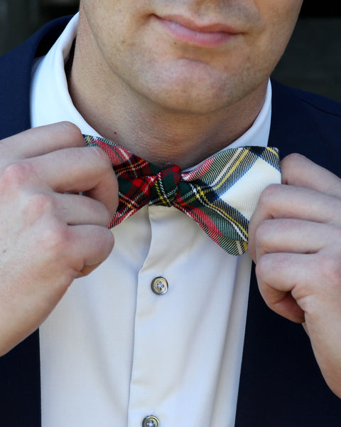 Person adjusting a plaid bow tie by Taylors Tartans with a white shirt and dark jacket.
