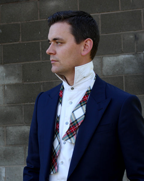 Man looking away from camera wearing a navy blazer, white shirt, and plaid untied tie against a brick wall.