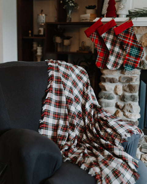 A red, white, and black plaid throw blanket with fringed edges is draped over a dark gray armchair in a cozy living room setting, with Christmas stockings hanging by a stone fireplace in the background.
