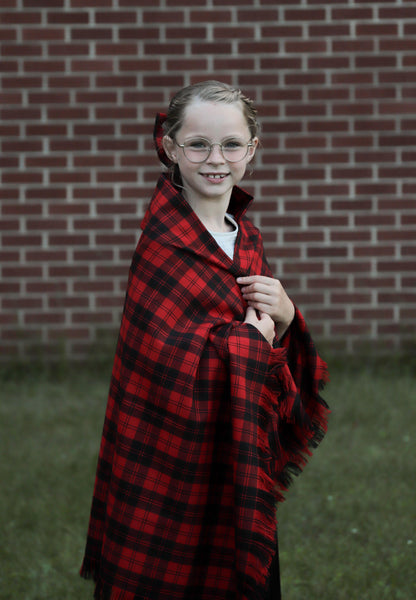 Young girl with long blond hair wearing an oversized red and black check scarf, standing in front of a brick wall