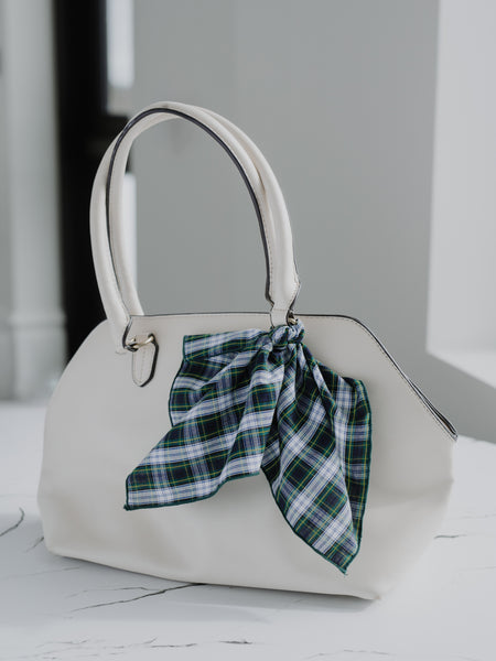 An off-white structured handbag with double top handles and a green plaid scarf tied in a bow on the front, set against a soft gray background.
