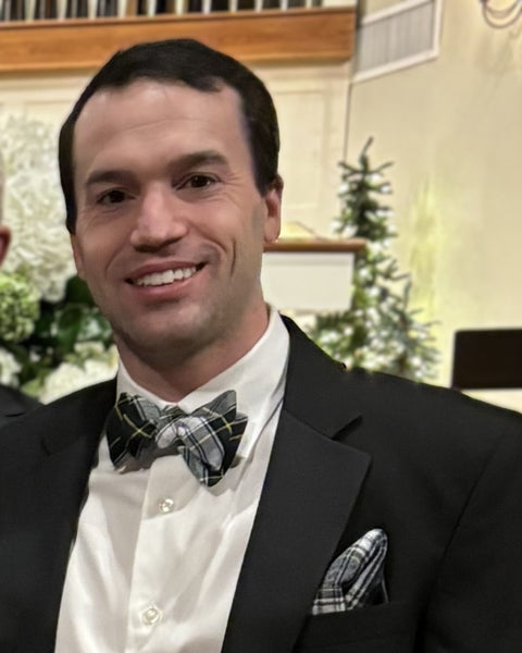 Man wearing a black suit with a green and white plaid bow tie and pocket square, indoors, at wedding.