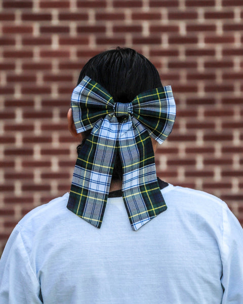 woman wearing a large navy and green plaid bow on their head against a brick wall.