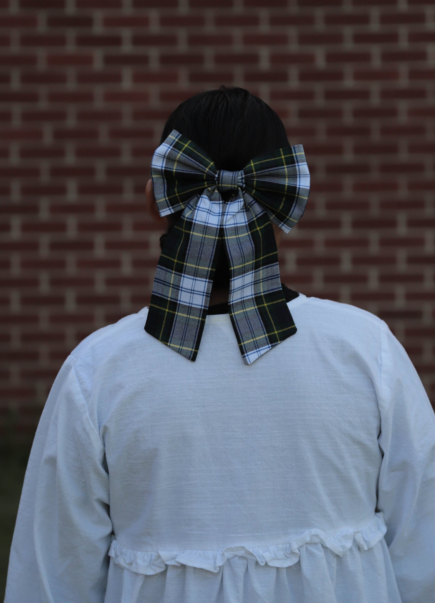 Woman wearing a large green, navy, white plaid bow in their hair against a brick wall.