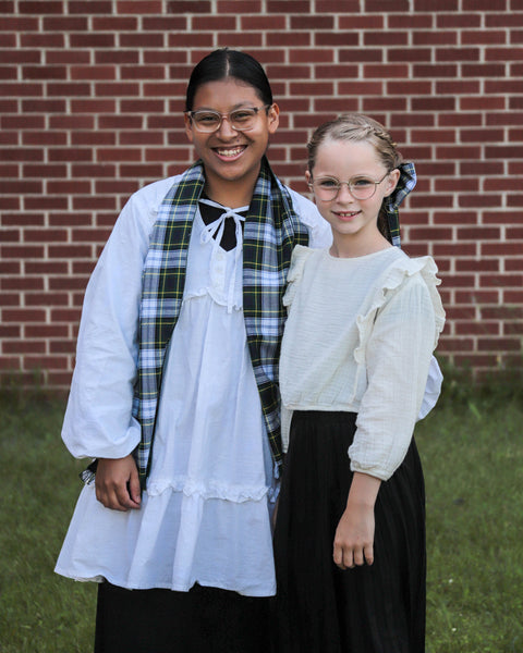 Two young girls wearing white blouses and navy and green scarf and hair bows standing in front of a brick wall.