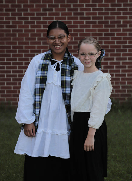 Two teenagers in white blouses wearing navy and green plaid scarves standing in front of a brick wall.