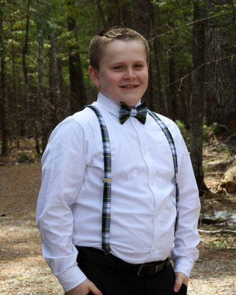 Young boy in formal attire with navy, green and white plaid suspenders and bow tie standing in a forest.