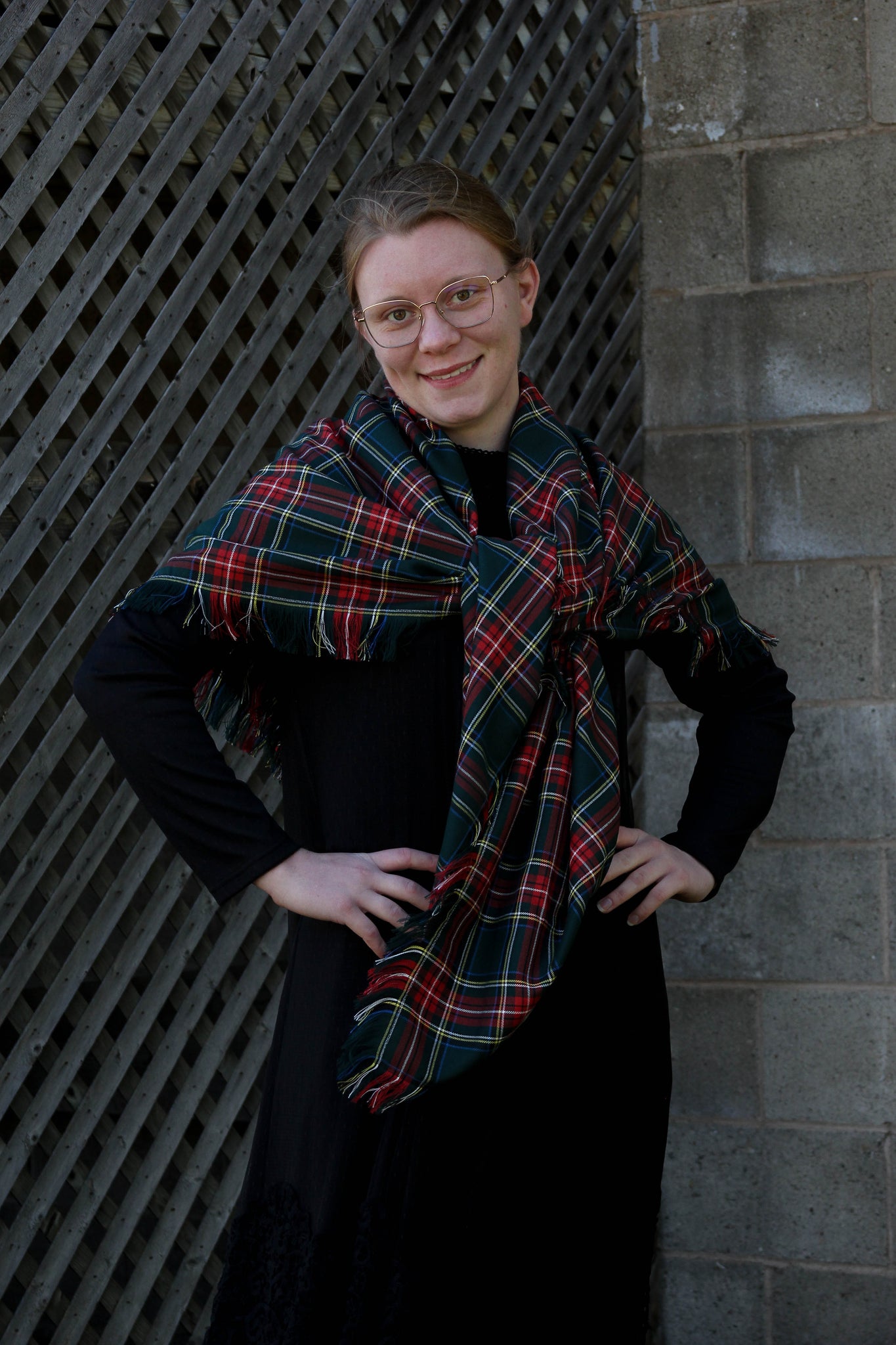 A woman standing with a Green Stewart green, red, and white plaid scarf draped over her shoulders, smiling against a backdrop of a grid-patterned wall.