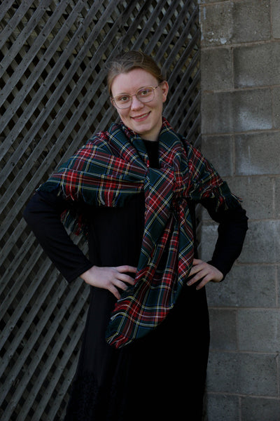 A woman standing with a Green Stewart green, red, and white plaid scarf draped over her shoulders, smiling against a backdrop of a grid-patterned wall.