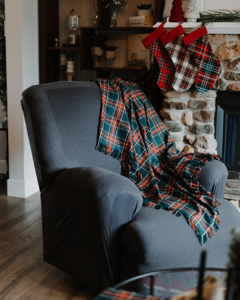 A dark gray upholstered recliner chair in a cozy living room, with a Green Stewart tartan plaid throw blanket draped over the back. The blanket has a tartan pattern in teal, red, orange, and white.
