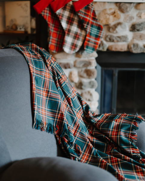 A plaid throw blanket draped over a gray armchair in a cozy living room setting, with a stone fireplace and red stockings visible in the background.
