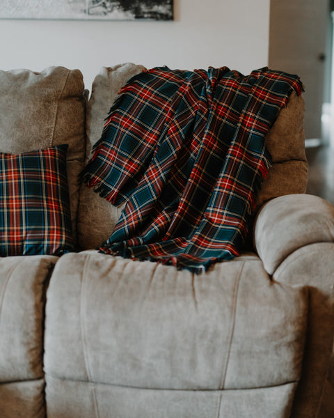 A red, green, and blue plaid Green Stewart tartan throw blanket with fringe is draped over a beige fabric recliner sofa, next to a matching plaid pillow.