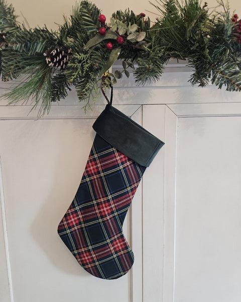 A Christmas stocking with a red, green, and yellow plaid pattern and a dark green cuff, hanging from a green pine garland on a white paneled wall.  Green Stewart tartan christmas stocking