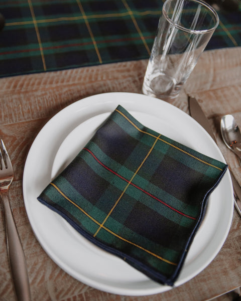 A Hunting Stewart tartan similar napkin in green, navy, and yellow tartan is displayed on a white plate, set on a rustic wooden table with a glass, silverware, and a matching plaid runner in the background.