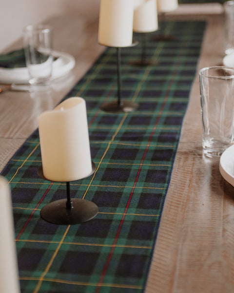 A green and navy plaid Hunting Stewart tartan similar table runner laid down on a wooden dining table, styled with black candlesticks holding white pillar candles and clear drinking glasses.