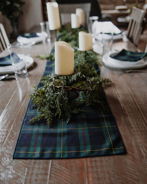 A green, navy, red and gold tartan plaid table runner styled on a rustic wooden dining table with white candles, greenery, and place settings.