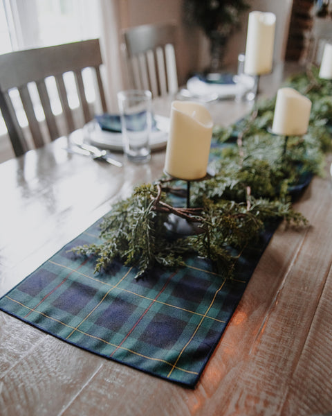 A green and navy plaid table runner with thin yellow and white stripes laid out on a rustic wooden dining table, decorated with evergreen branches and cream pillar candles.