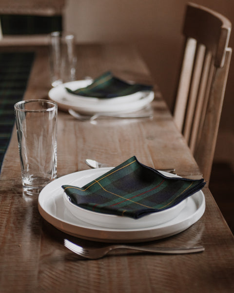A close-up of a place setting on a rustic wooden dining table, featuring a folded Hunting Stewart tartan similar plaid napkin in dark green, navy, and yellow over white stacked plates, with a clear glass and fork nearby.