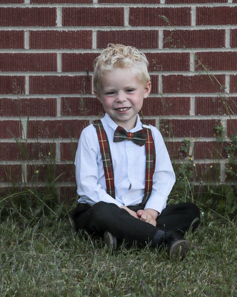 Young boy wearing a white shirt, black pants and a Prince Edward Island tartan bow tie and suspenders  sitting on grass against a brick wall