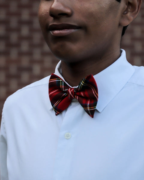 Person wearing a white shirt with a red plaid Royal Stewart tartan bow tie against a brick wall.