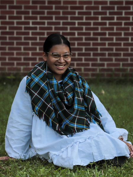Girl wearing a white outfit with a dark green and blue plaid shawl sitting on grass against a brick wall.