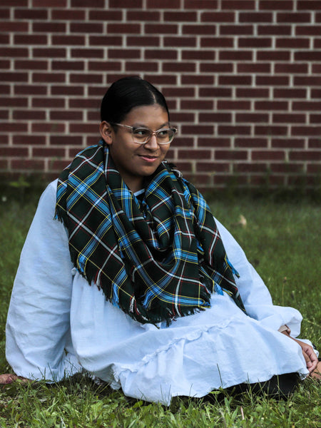 Person wearing a white outfit and plaid scarf sitting on grass against a brick wall.