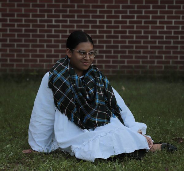 Woman sitting on grass wearing a white outfit and Labrador tartan scarf against a brick wall.