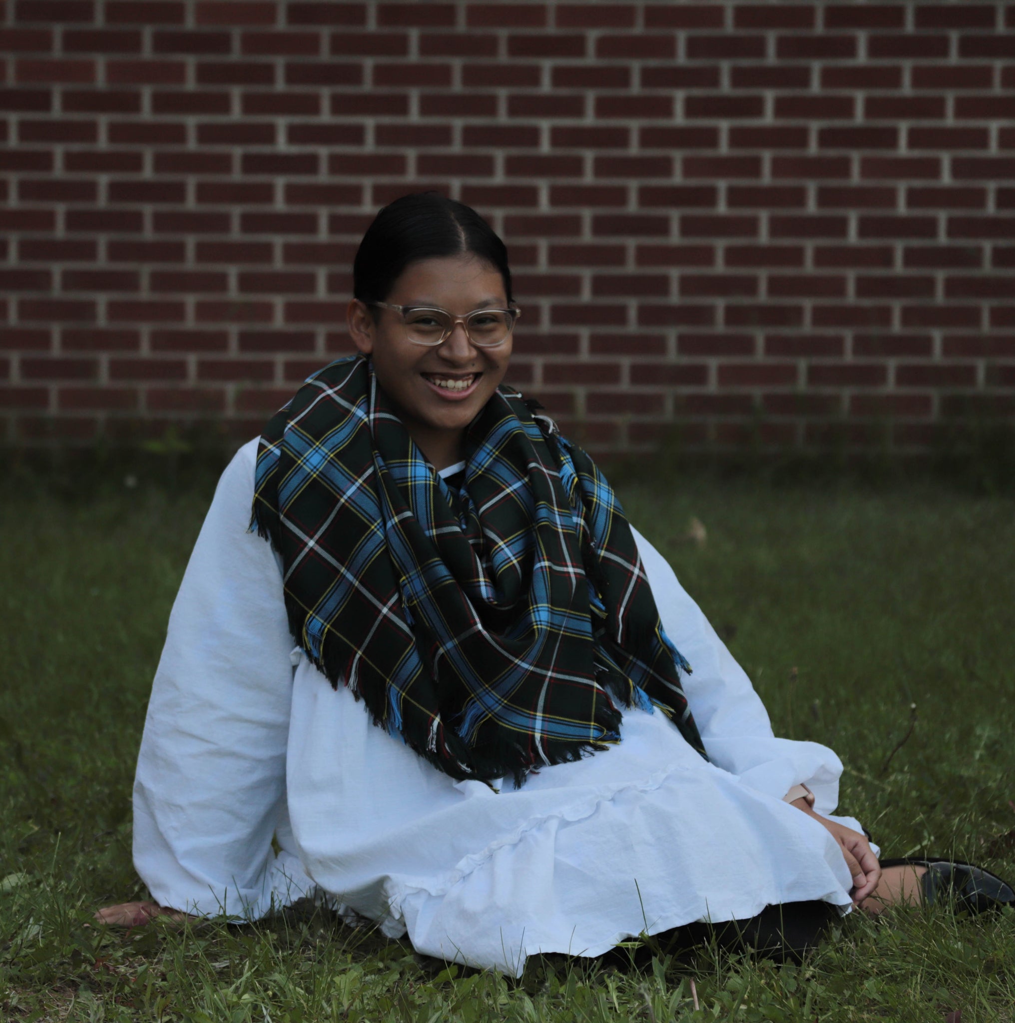 Person wearing a white dress and Labrador plaid blanket scarf sitting on grass with a brick wall background