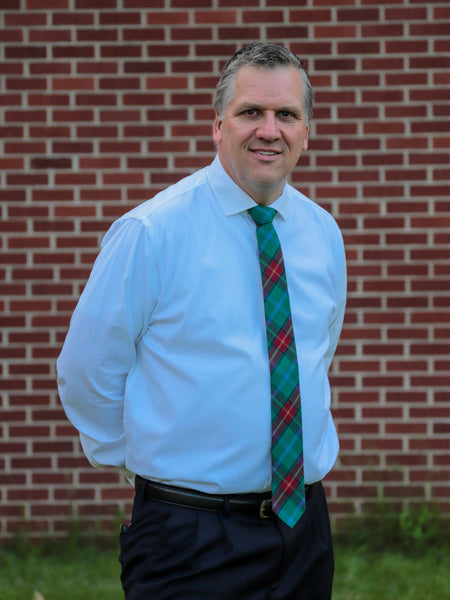 Man wearing a light blue shirt and Manitoba plaid tie standing in front of a brick wall.