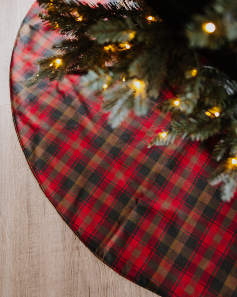 A round Christmas tree skirt with a red, black, and brown plaid pattern, shown under a decorated Christmas tree with warm lights on a light wood floor.