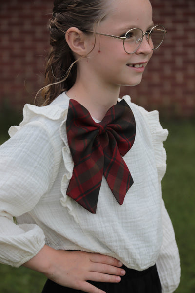 Young girl wearing a white blouse with a large red plaid bow, standing outdoors.