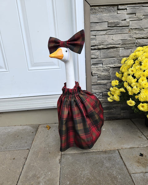 White porch goose wearing Maple Leaf tartan dress and bow in front of white door with brick wall and yellow mums