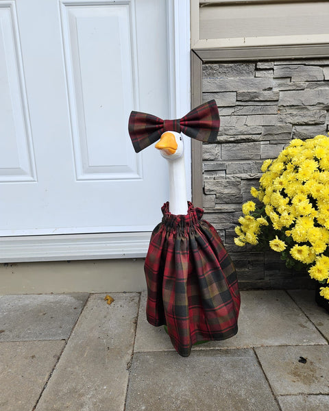 Cement goose wearing plaid dress in front of white door, brick wall, Yellow Mums plant, standing on cement blocks