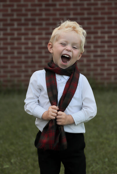 Laughing child wearing a white shirt and a brown, red, and gold plaid scarf, standing in front of a brick wall.