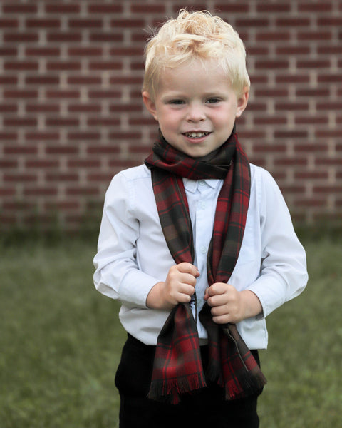 Young boy wearing a red green gold and brown plaid scarf in front of a brick wall.