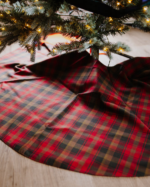 A red, brown and gold plaid Christmas tree skirt spread on a light wood floor beneath the branches of a decorated Christmas tree with warm string lights.