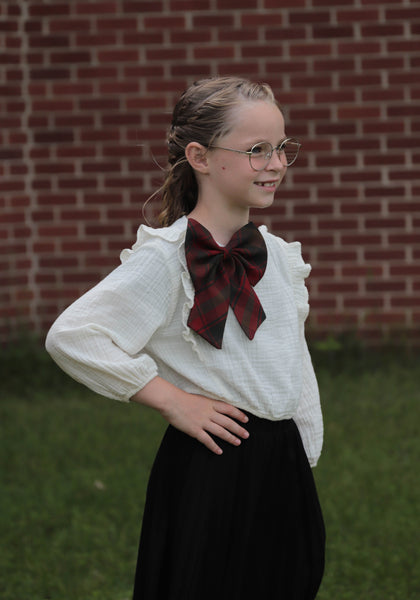 Girl with long blond hair, wearing white blouse, and brown and red plaid bow on front of her blouse.  
