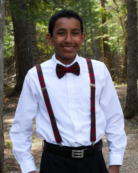 Young boy wearing a white shirt, brown and red plaid suspenders, and  bow tie standing in a forest.