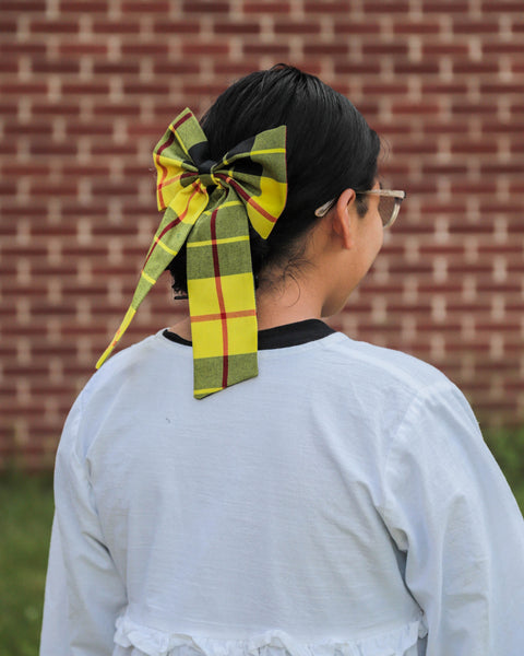 Woman with a large yellow and black plaid bow in their hair against a brick wall.