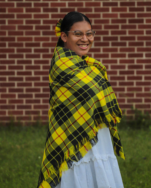 Person wearing a yellow and black plaid blanket over shoulders against a brick wall.