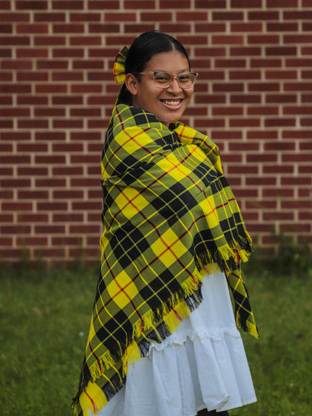 woman wearing a bright yellow and black plaid shawl in front of a brick wall.