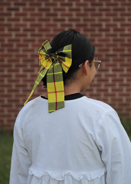 Person wearing a large yellow plaid bow in their hair against a brick wall.