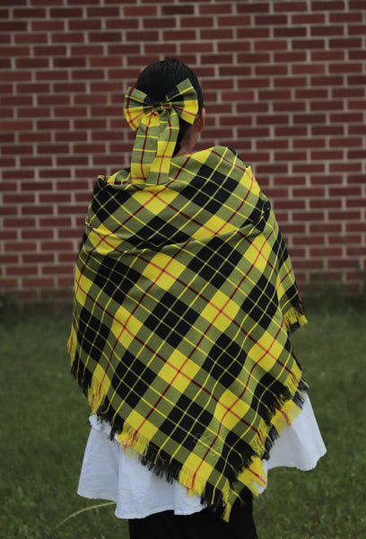 Person wearing a yellow and black Mcloed plaid shawl in front of a brick wall.