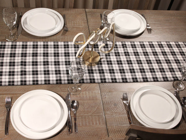 Dining table set with white plates, silverware, and a black and white checkered table runner, and a brass sign that says "joy".